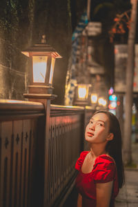 Portrait of young woman looking away while standing against illuminated wall