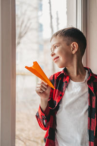 A boy with an orange paper airplane, sits at the window and looks out into the street. boy dreams 