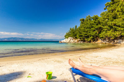 Low section of woman relaxing at beach against blue sky