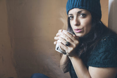 Portrait of woman blowing hot coffee while sitting by wall