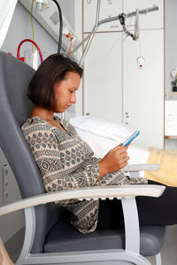 Side view of young woman using digital tablet while sitting on chair at hospital