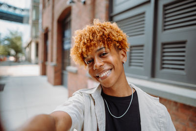 Happy young man in hat and eyewear taking a selfie near building with red hair