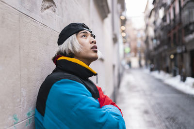 Thoughtful male hipster looking up against wall in city during winter