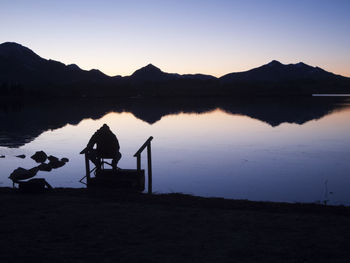 Silhouette man sitting by lake against sky during sunset