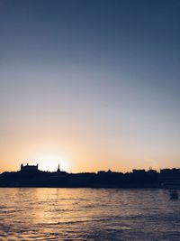 Silhouette buildings by sea against clear sky during sunset