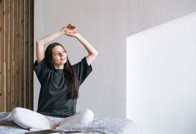 Young smiling woman teenager girl with dark long hair in jeans sitting on bed in her room at home