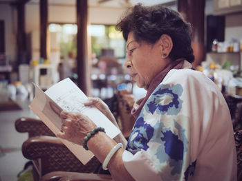 Side view of woman reading book while sitting on chair at home