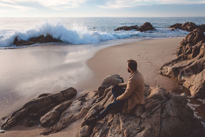 Side view of man sitting on rock at beach against sky