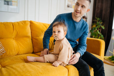 Child with a pacifier sitting on a yellow sofa with his father