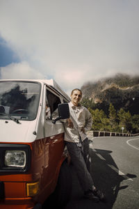 Portrait of smiling young man standing next to van on road during road trip