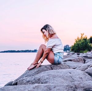 Side view of man sitting on rock against sky during sunset