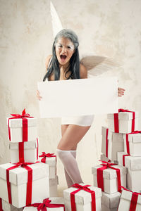 Angry female model in angel costume holding blank cardboard amidst christmas presents