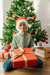 Portrait of a boy in a santa claus hat with a huge gift box in his hands sits on the floor 