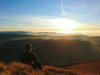 Rear view of hiker sitting on grass looking at sunset over mountains