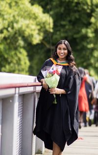 Portrait of young woman wearing graduation gown while standing on footpath against trees