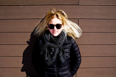 Portrait of young woman wearing sunglasses and warm clothing while standing against wall