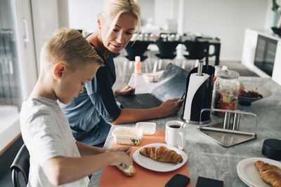Mature woman with laptop looking at son applying butter on croissant in kitchen