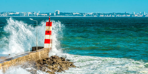 Waves splashing lighthouse by sea against clear blue sky