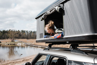 Girl looking out of roof top tent at a view of a lake whilst camping