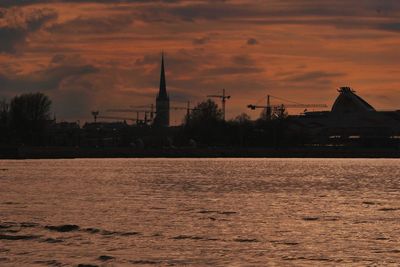 Silhouette buildings by river against sky at sunset