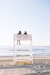 Rear view of couple sitting on lifeguard hut on sand at beach against sky