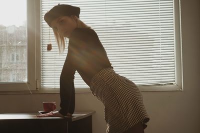 Side view of young woman standing by window at home