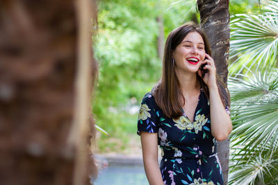 Beautiful young woman using phone while standing against tree trunk