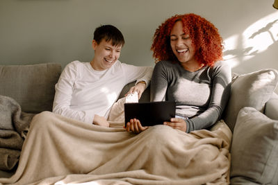 Cheerful young women sharing laptop while watching movie together sitting on sofa at home