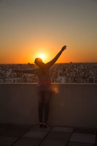 Rear view of woman standing by cityscape against sky during sunset