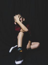 Boy covering face while sitting against black background
