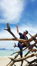 Young man pointing finger towards sky while sitting on driftwood at beach