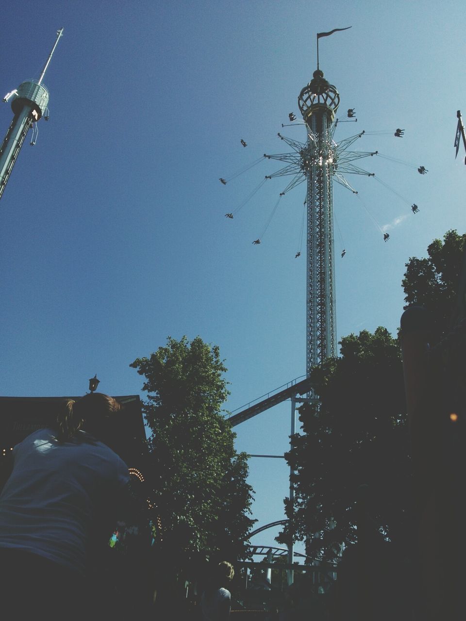 clear sky, low angle view, tree, sky, amusement park, tall - high, blue, built structure, technology, leisure activity, men, street light, building exterior, architecture, amusement park ride, arts culture and entertainment, transportation, copy space, outdoors