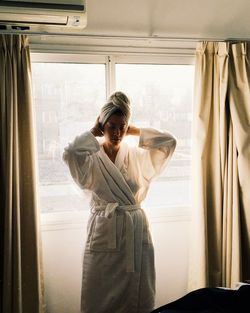 Woman wearing bathrobe while standing by window at home