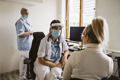 Female healthcare worker wearing mask and face shield talking patient in medical clinic