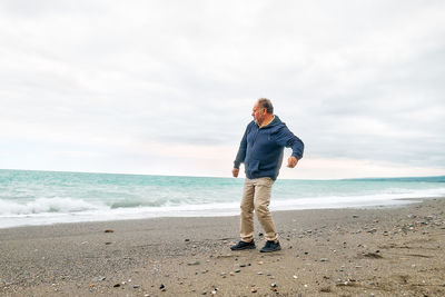 Happy middle-aged bearded man throws pebbles into the sea, walking along deserted winter beach. 