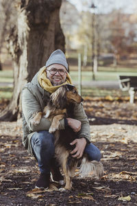 Portrait of smiling man embracing dog while crouching at park during autumn