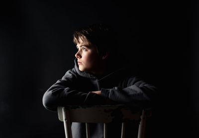 Low key portrait of adolescent boy sitting on a chair in a dark room.