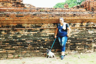 Full length portrait of woman with pug standing by brick wall