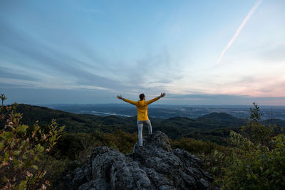 Rear view of man with arms outstretched standing on rock against sky