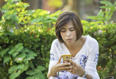 Woman using phone while sitting on seat against plants