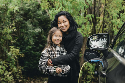 Portrait of mid adult woman with daughter by electric car at charging station