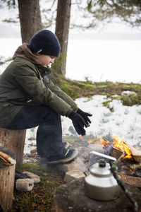 Side view of boy warming hands while sitting by bonfire in winter