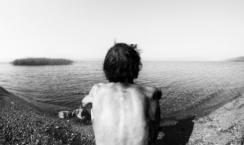 Rear view of shirtless man sitting at beach against clear sky