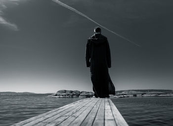 Rear view of man standing on pier at sea against sky
