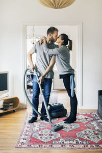 Full length of man kissing woman while vacuuming carpet at home