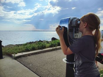 Woman looking through binoculars at lake against sky