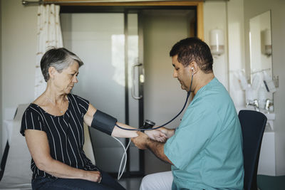 Male doctor checking blood pressure of senior woman while sitting in hospital