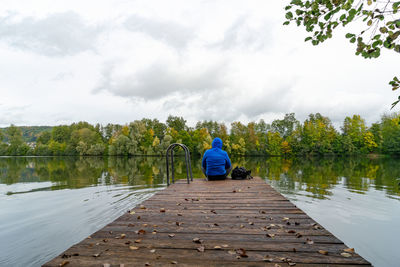 Rear view of man sitting on pier over lake against sky