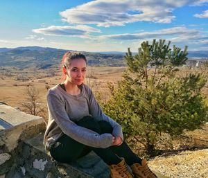 Portrait of woman sitting on retaining wall against sky during sunny day