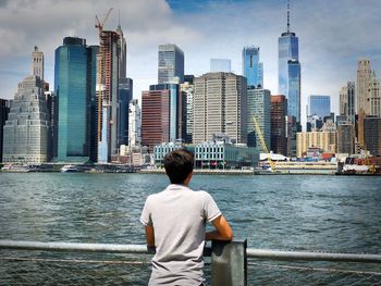 Rear view of teenage boy looking at river against skyscrapers in city
